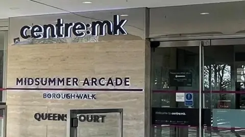 The outside of an entrance to Centre:MK, a shopping centre in Milton Keynes. There is a sign which says, centre:mk MIDSUMMER ARCADE BOROUGH WALK.