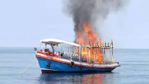 Tall orange flames rise into the sky on a small red, white and blue boat in the water off Koh Tao