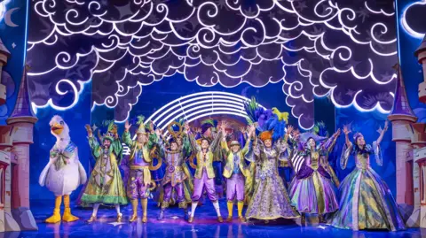 Paul Coltas Dozens of performers on the stage have bright smiles on their faces, and their hands raised. They are wearing costumes in shades of gold and purple. The set design depicts a cloudy sky above them. There is a person in a goose costume on the left.