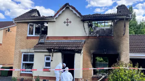 Forensic officers wearing white suits are outside a house that has been destroyed in a fire. It is a burnt-out shell with views through to the back garden. The forensic officers are working where the front door would have been. Red and white fire tape is outside. 