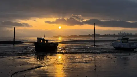 WeatherWatchers/Sparky A stretch of wet sand leading to the sea just as the sun begins to rise over the water. The scene is still dark with ominous clouds in the sky, and a boat on the left is silhouetted by the sun behind. There is another white boat to the right. Both are moored by the shore