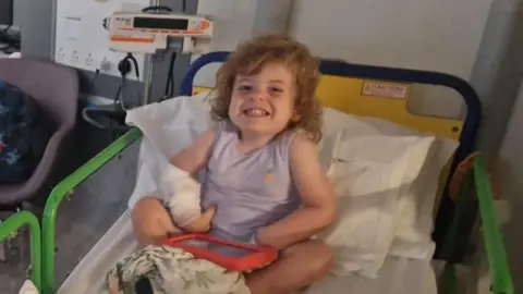 Contributed Wilfred is pictured smiling to camera while sitting in a hospital bed. He has a white bandage on one arm. A children's tablet can be seen in his lap along with a blanket. He is wearing a purple vest top. Hospital equipment sits next to the bed.