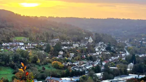BBC Weather Watchers/Quatro The sun rises above the wooded Stroud Valleys, painting the sky a golden hue. There are lots of houses in the valley, along with a major road. The trees are different shades of green, yellow, red and brown.