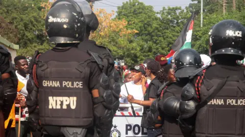 Three police officers wearing black helmets and protective jackets with the words "Ghana Police NFPU" . They are facing a crowd of people some holding flags.