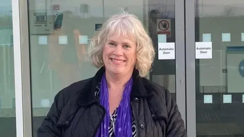 A woman with white, wavy hair who is smiling facing the camera. She is wearing a dark blue coat, blue scarf and black and white top. There are glass automatic doors behind her.