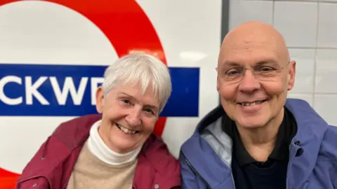 Woman on left has short cropped white hair, pink jacket and beige jumper. She is sat next to a man who is bald, wears a blue jacket and black t-shirt. They are both smiling and sat in front of a Stockwell Tube roundel sign.