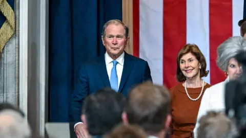 Reuters Image shows former US President George W Bush, wearing a blue suit and lighter blue tie, standing next to his wife Laura Bush. They are in front of a large Stars and Stripes flag. 