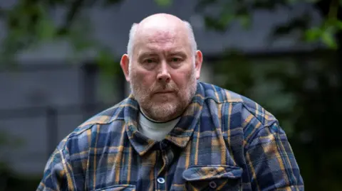 PA Media A man in a plaid shirt with a beard looking ahead with a neutral facial expression.