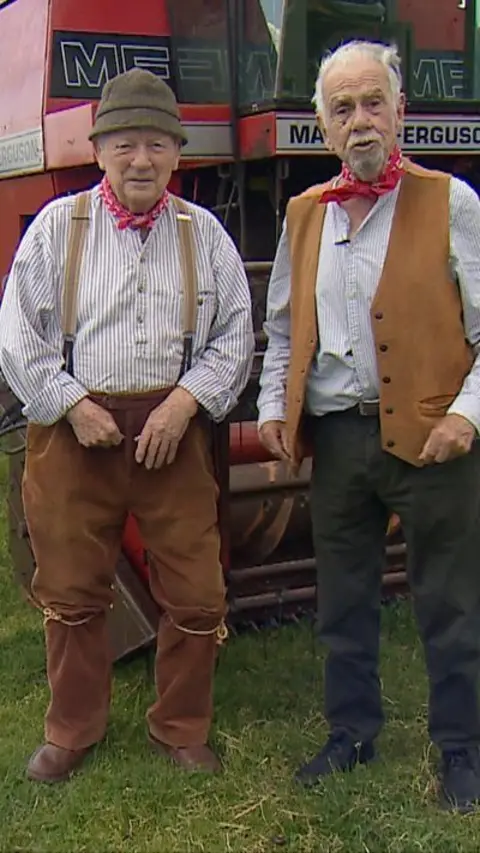 Tommy and Pete wear red neckerchiefs, stripy shirts, and dark trousers. They are standing in a field in front of a combine harvester. 