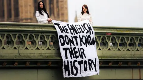 Jude Harrison photograph of two women standing on a bridge outside Westminster beside a large banner draped over the railing. The banner reads, “THE ENGLISH DON’T KNOW THEIR HISTORY” in bold, hand-painted capital letters. 