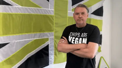 Dale Vince in a black t-shirt stood in front of a green, black and white union flag with his arms crossed. 