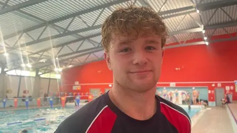 A young man has wet, curly light brown hair. He is standing near a swimming pool. He is wearing a black and red swimming club t-shirt