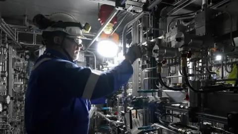 A man in a blue boiler suit wearing a hard hat stands in front of machinery.
