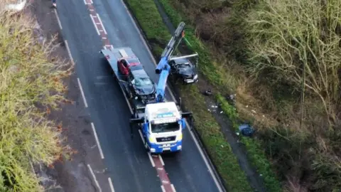 An aerial image of a lorry parked in the middle of a road, which has trees either side of it. A crane attached to the lorry is lifting a damaged black BMW out of a ditch. The lorry already has two damaged cars loaded onto its rear shelf.