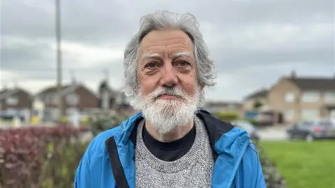 BBC A man with a beard in a grey jumper and blue jacket, with houses and grass in the background
