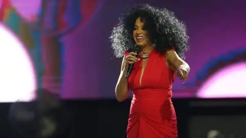 Diana Ross on stage wearing a red dress, smiling and pointing towards the audience