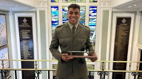 Taylor Lawrence Taylor Lawrence, smiling in a military uniform, stands in front of a stained glass window, with wooden mounted lists of names either side of him holding a small metal trophy-style knife.