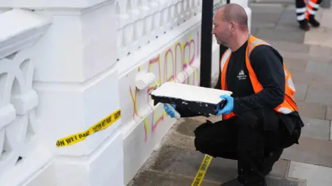 Eddie Mitchell A man crouches down and paints white over the words "tax evasion" wrote in pink on a white wall