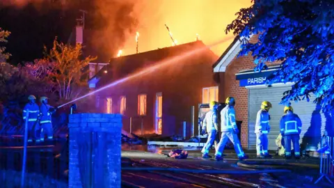 PACEMAKER Firefighters fighting a blaze at the church in Greenisland last weekend. One firefighter is trying to extinguish the fire with a hose. The windows are red with the fire and smoke is in the air.