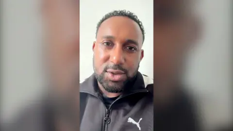 A man with short black hair, moustache and beard talks to the camera. He is wearing a zipped up sports jacket.