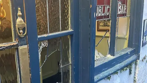 An exterior view of smashed glass panes on the windows and door of The Boot Inn. A sign on blue-framed windows says 'Public Bar'.