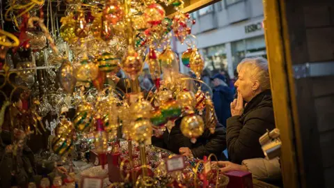 Getty Images A close-up view of a Christmas market stall, overflowing with ornate holiday decorations and bathed in warm, golden light. In the foreground, dozens of delicate glass ornaments hang densely from wooden beams. On the right side, an elderly woman in a dark winter coat stands thoughtfully at the stall.