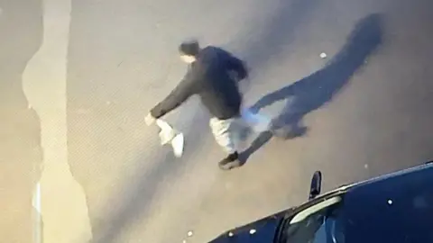 A slightly blurred CCTV image showing a man wearing black trainers, grey joggers and a black coat holding a pigeon by one wing.
