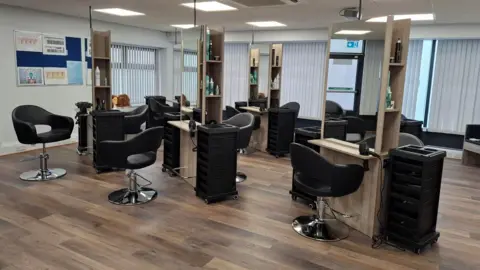 Handout The fully kitted out hairdressing salon can be seen with numerous large mirrors, client seats and tool trollies. The large room has medium brown laminate wood flooring and large windows covering with vertical blinds.