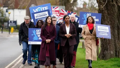 PA Media Kemi Badenoch in a brown suit and white top walking down a street with a small crowd of activists holding blue placards reading "stronger economy, stronger country".