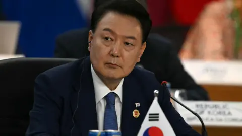 Getty Images South Korean President Yoon Seok-yeol sits in a meeting in a dark blue suit behind the South Korean flag