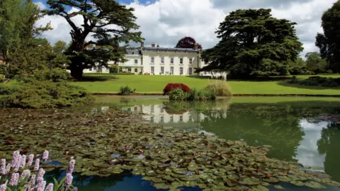 Kings West Estate Agents The back façade of Moulton Grange is in the centre of the picture, framed by mature trees, with a wide lawn and a lake with lilies and ornamental plants in the foreground.