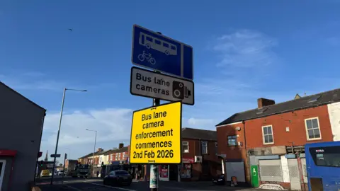 An image showing three signs, the top is a blue bus lane sign, with a white image of a bus and a bicycle, below that, a white sign with black letters and an image of a camera, then below that, a yellow sign with black letters saying, bus lane camera enforcement commences 2nd Feb 2026. The signs are on New Hall Lane in Preston, and in the background you can see boarded up shops in red bricked terraces. The sky is blue.