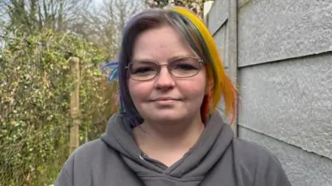 A woman with rainbow coloured hair stands next to a white fence in an alleyway. She is wearing a grey hoodie and is wearing glasses. On the left of the photo is a chain link fence covered in ivy. 