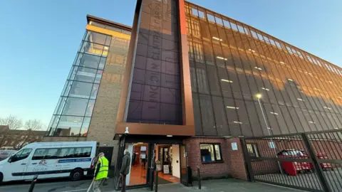 A large, modern multi-story car park, designed with a frontage made up of grills and black-framed glass panels. A white van is parked outside and two men can be seen walking towards it.