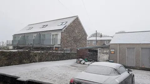 Wolf Fleece/BBC Weather Watchers Camborne in Cornwall on 5 January. A snowy scene. There is a parked car that is frozen over. There are three buildings, two in the foreground and one in the background, both have snow on the roofs. The pavements are snowy. The sky is grey. 