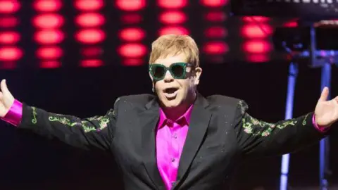 Getty Images Elton John stands on stage during a show in 2017 in Monaco. He wears a black suit with a bright purple shirt underneath as well as sunglasses. He has his arms outstretched either side of him.