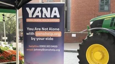 YANA A sign is standing just off centre to the left. It has a purple background, and says YANA in big white writing. On the right is the front of a green tractor with a yellow wheel.  