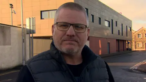 Councillor Paul Dunlop is standing on a street. He wears glasses and has a navy body warmer on.