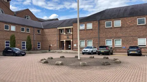 Grey roofed, red brick building overlooking a court yard / parking area. There is an off-white flag pole in the centre. Four cars are parked around the edge of the parking area.