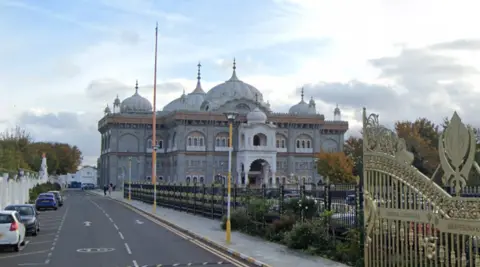 A Google Street image of a Gurdwara. The large, white building has domes on its roof and is beyond gold gates.