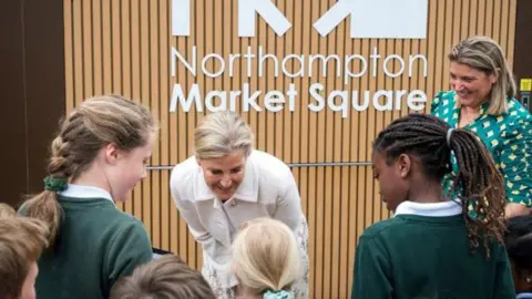 West Northamptonshire Council Blonde haired woman crouching down smiling and looking at five students wearing green school sweatshirts. They are standing in front of a wood paneled wall with white writing 'Northampton market square' written across it.