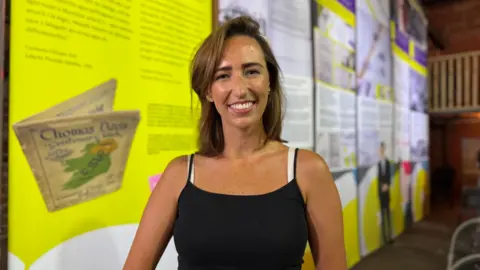 A young woman with shoulder-length blonde hair smiles broadly at the camera. She is wearing a black top and is standing in front of tall exhibition displays with text and images.