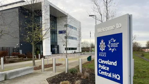 A large white and grey multi-storey building in the background. In the foreground is a Welcome to Cleveland Police Central HQ sign.