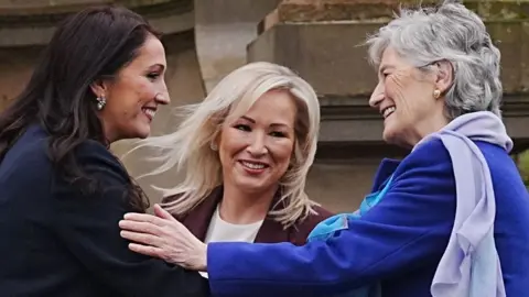 Ireland's President Catherine Connolly, with short grey hair, wearing a blue coat, shaking hands with Deputy First Minister Emma Little-Pengelly, who has long brown hair and wearing a navy jacket. First Minister Michelle O'Neill, with long blonde hair, is standing between them, smiling.