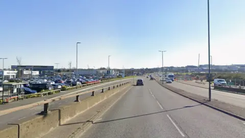 A dual carriageway running through an industrial part of the city. There are some cars and lorries on the road. It is a sunny day. 