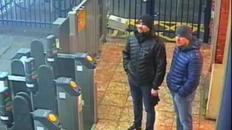 Two men in dark coats and hats beside rail station ticket barriers.