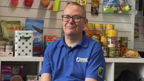 A close-up a man wearing glasses and a blue shirt. He has cropped hair aand is smiling as he sits in front of shelves stacked with pet food.