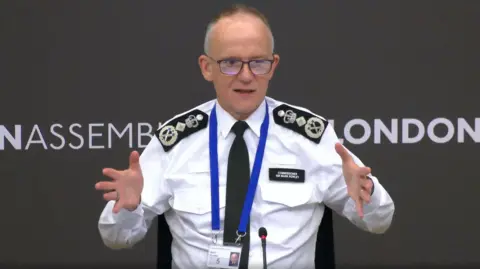 London Assembly Sir Mark Rowley addresses the Police and Crime Committee. He is wearing a white shirt with a black tie and there is a lanyard around his neck.