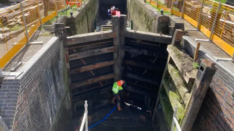 Image shows two brand new lock gates closed in place after being replaced. There is a workman standing with his back to camera as if he's about to push the lock gates open to test them. He is wearing a yellow florescent jacket and bright orange helmet and black trousers. Beyond the gates is the next set of gates further down, and there is no water currently in this part of the canal. There are old 200-year-old walls either side of the gates and rest of canal. In ground level above the gates, is yellow metal fencing where the rest of the equipment is stored. 