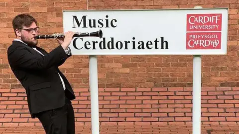 Ianto Williams pictured outside Cardiff's school of music. He is playing a clarinet, and wearing a black suit. 